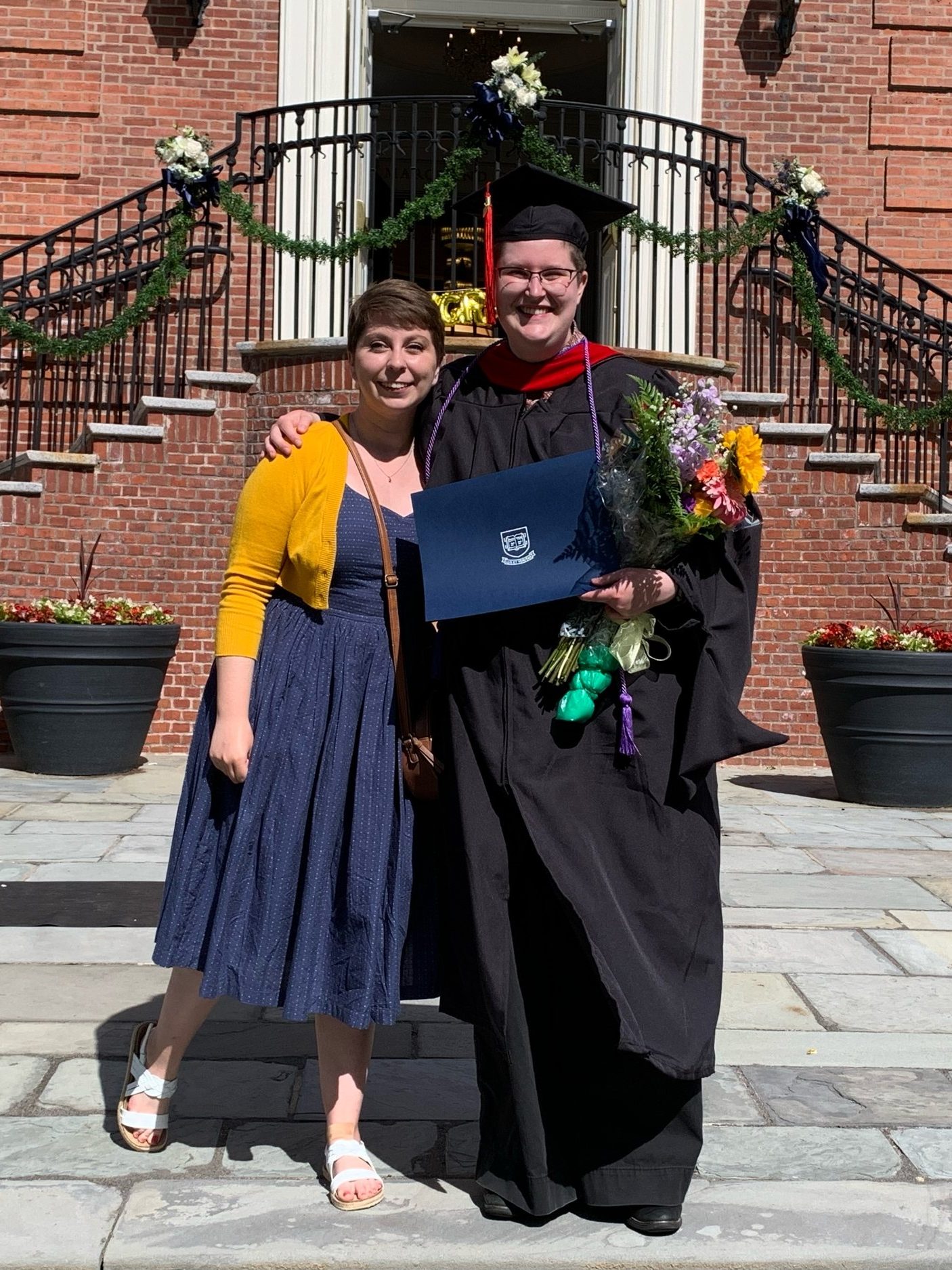 Two white people standing smiling in the quad of Yale Divinity School. The person on the right is wearing full Master's degree regalia, having just graduated.